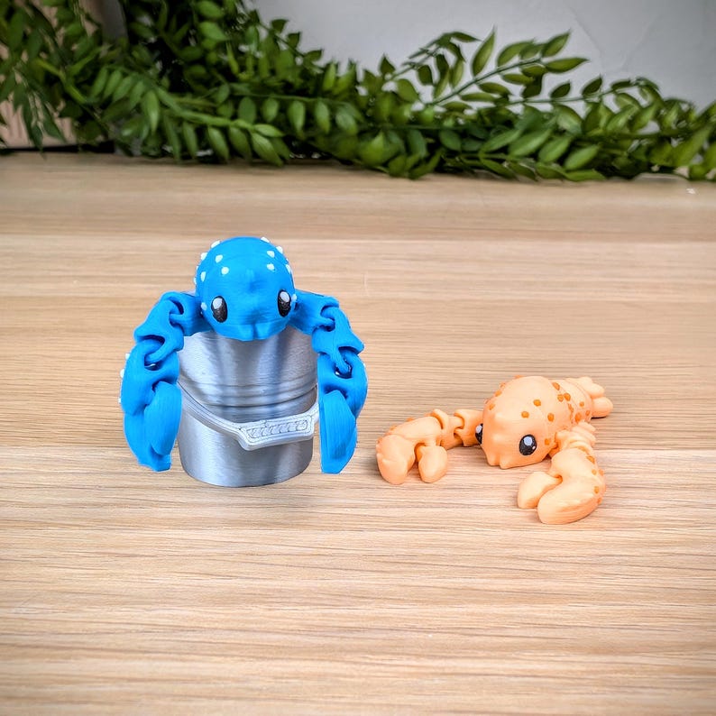 May include: Two small, colourful, 3D printed lobster figurines. One is blue with white spots and is holding a small silver bucket. The other is orange with white spots.