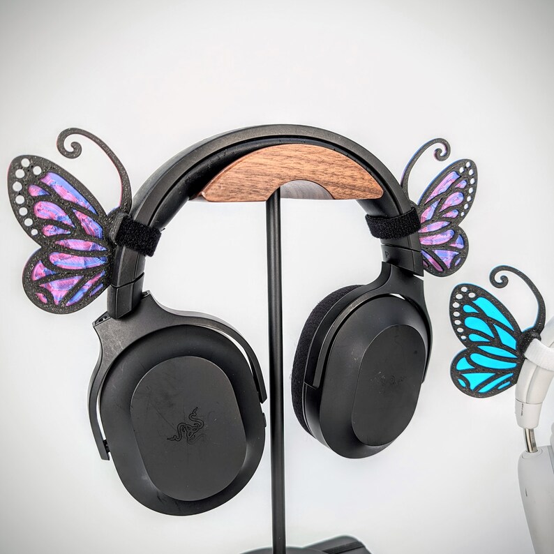 May include: Black gaming headset with a wooden stand and butterfly-shaped decorations. The butterflies are pink, blue, and purple.
