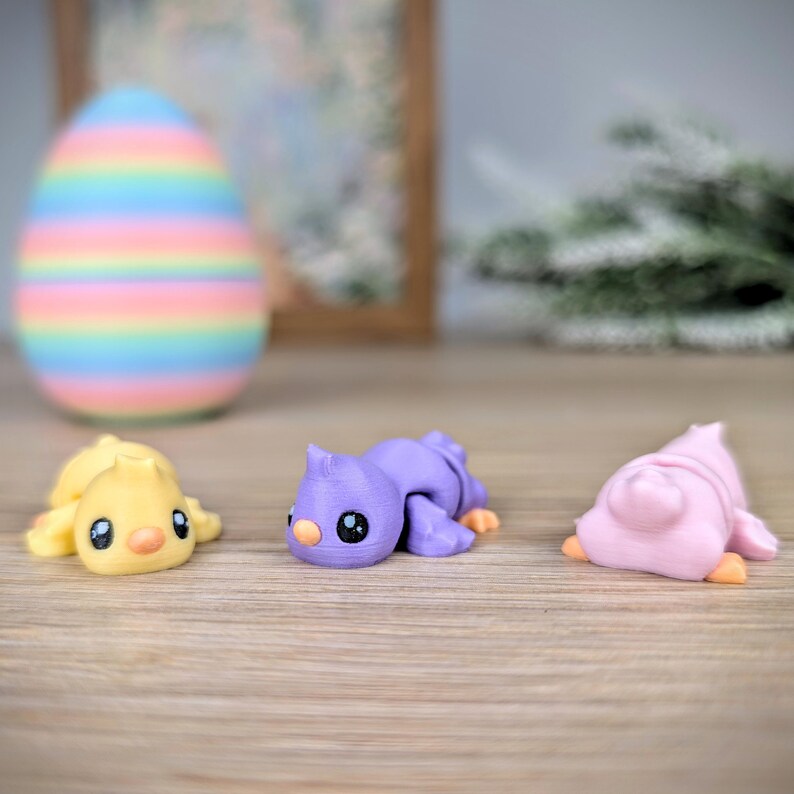 May include: Three colorful, 3D-printed animal figurines: a yellow chick, a purple bird, and a pink bird. A large, pastel-striped Easter egg is in the background. The figurines are lying down on a wooden surface.