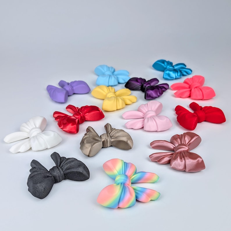 May include: A collection of colorful bow-shaped accessories in various shades, including red, pink, blue, purple, and yellow. The bows are arranged on a white surface, with a rainbow-colored bow in the foreground. The bows appear to be made of a smooth, possibly satin-like material.