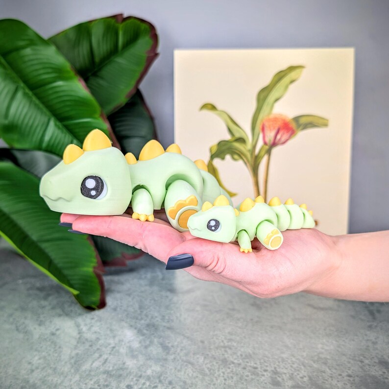May include: Two articulated green and yellow toy dinosaurs, one large and one small, are displayed. The larger dinosaur is in the foreground, with the smaller one behind it. The dinosaurs have large eyes and yellow accents. A painting of a flower is in the background.