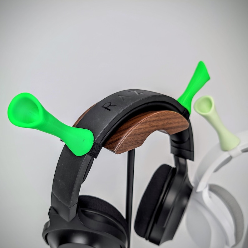 May include: Headphone stand with a wooden arch and green accents. The stand holds a black Razer headset. The stand has a unique design with green spoon-shaped holders. A white headset is also visible.