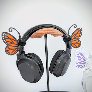 Puede incluir: Auriculares negros con un soporte de madera y dos decoraciones en forma de mariposa. Una mariposa es naranja y la otra es morada.