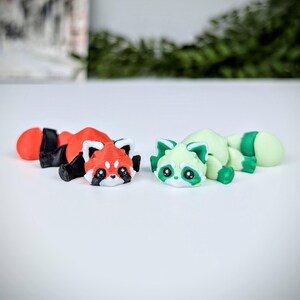 May include: Two small, hand-painted figurines of red pandas. One is red with black markings and the other is green with white markings. Both have black eyes and are lying on their sides.