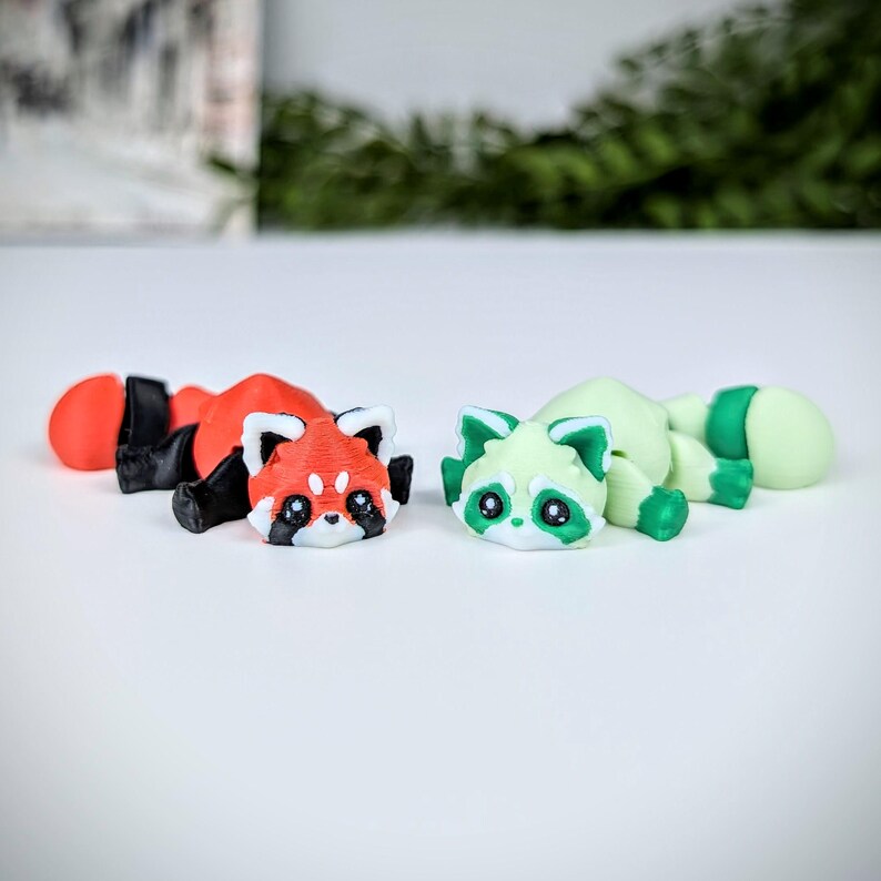 May include: Two small, hand-painted figurines of red pandas. One is red with black markings and the other is green with white markings. Both have black eyes and are lying on their sides.