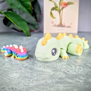 May include: Two colorful, cartoon-style dragon figurines. One is a rainbow of colors, the other is light green with yellow accents. Both have large, expressive eyes and are lying down. The background includes a botanical print and green leaves.