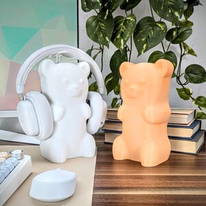 May include: Two gummy bear shaped night lights, one white and one orange, on a wooden surface. The white gummy bear is wearing headphones.