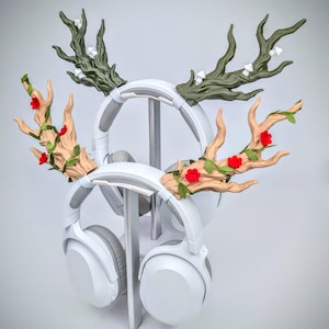 May include: White headphones with decorative antler-shaped attachments. The antlers are painted in shades of brown and green, adorned with red flowers and green leaves. The headphones are displayed on a silver stand.