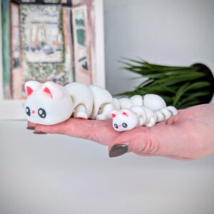 May include: Two white cat-shaped figurines with segmented bodies, resembling caterpillars. The larger cat has pink inner ears and red feet, while the smaller one mirrors the design. Both have black eyes and are held in a hand.