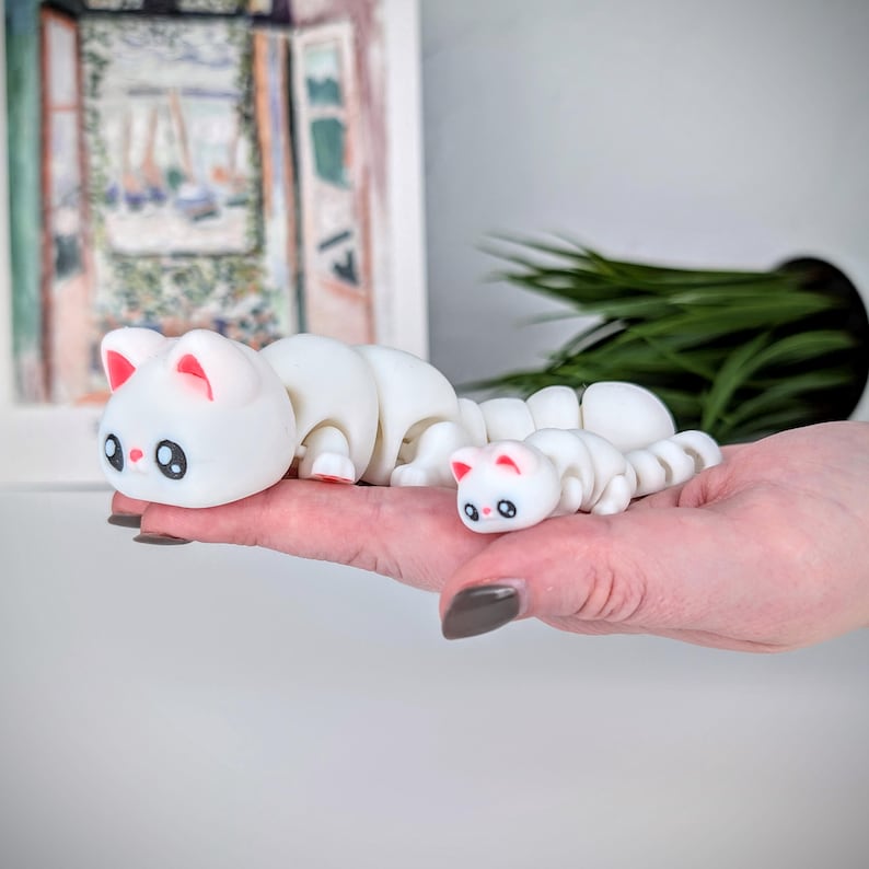 May include: Two white cat-shaped figurines with segmented bodies, resembling caterpillars. The larger cat has pink inner ears and red feet, while the smaller one mirrors the design. Both have black eyes and are held in a hand.