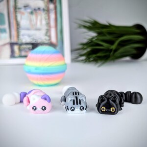 May include: Three small cat figurines in pink, gray, and black. The pink cat has purple ears and a white tail. The gray cat has black stripes and a white face. The black cat has yellow eyes. A rainbow-colored ball is in the background.