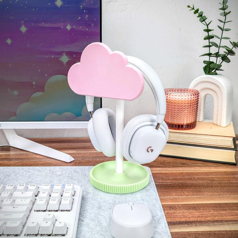 May include: A pink cloud-shaped headphone stand with a white base and green accents. The stand is holding a pair of white headphones with a black logo. The stand is on a desk with a keyboard and a mouse.