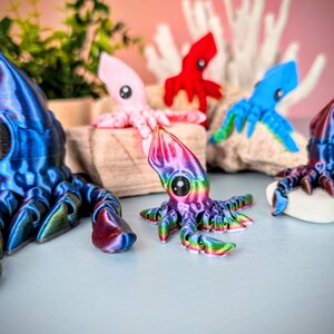 May include: A collection of colorful, 3D printed squid figurines. The squid are in various poses and have iridescent finishes. The squid are displayed on a wooden surface with a coral background.