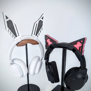 Pode incluir: Dois conjuntos de fones de ouvido em suportes pretos. Um &eacute; branco com acess&oacute;rios de orelhas de coelho, o outro &eacute; preto com acess&oacute;rios de orelhas de gato. As orelhas de coelho s&atilde;o brancas com detalhes em preto, e as orelhas de gato s&atilde;o pretas com detalhes em rosa.