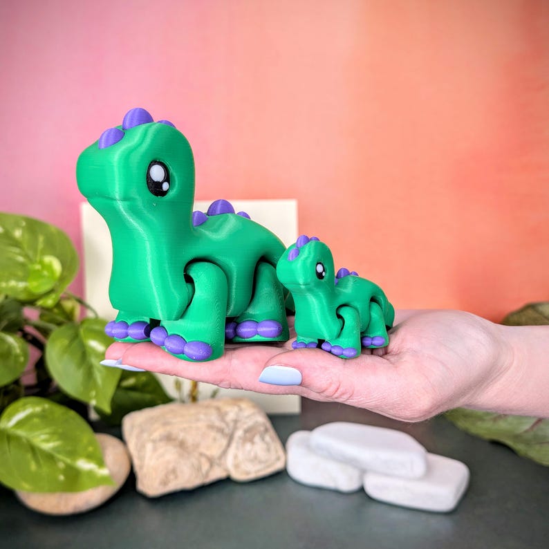 Op de afbeelding: Twee groene dinosaurusbeeldjes met paarse accenten. De grotere dinosaurus staat voor een kleinere. De dinosaurussen hebben grote, ronde ogen en worden op een hand gehouden. De achtergrond is een verloop van roze en oranje.