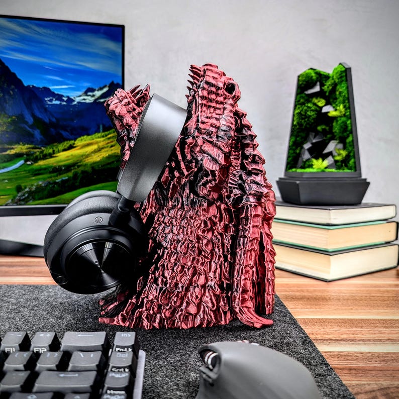May include: A red, textured, dragon-shaped headphone stand with a black headset resting on it. The stand is on a gray surface with a black keyboard and a black mouse.