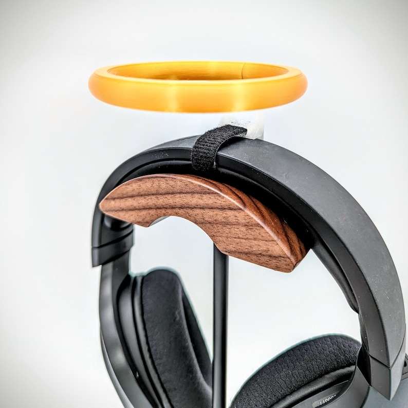 Op de afbeelding: Een zwarte koptelefoonstandaard met een accent van walnoot hout en een gele ring bovenop. De standaard is ontworpen om koptelefoons veilig te houden.
