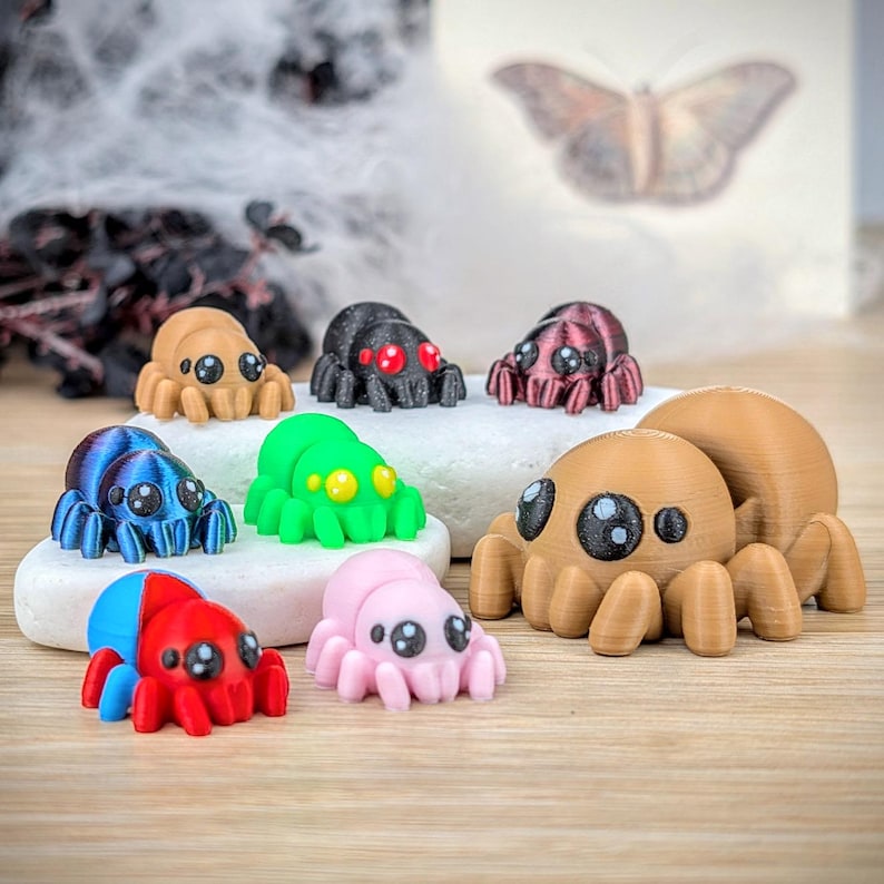 May include: A collection of colorful, 3D-printed spider figurines in various colors, including brown, black, blue, green, pink, and red. Each spider has large, cartoonish eyes. The spiders are displayed on a white stone and wooden surface.
