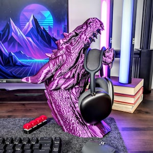 May include: A purple dragon head-shaped headphone stand. The stand is 3D-printed and holds a pair of black headphones. The dragon has detailed scales and a menacing expression. The stand sits on a gray surface.