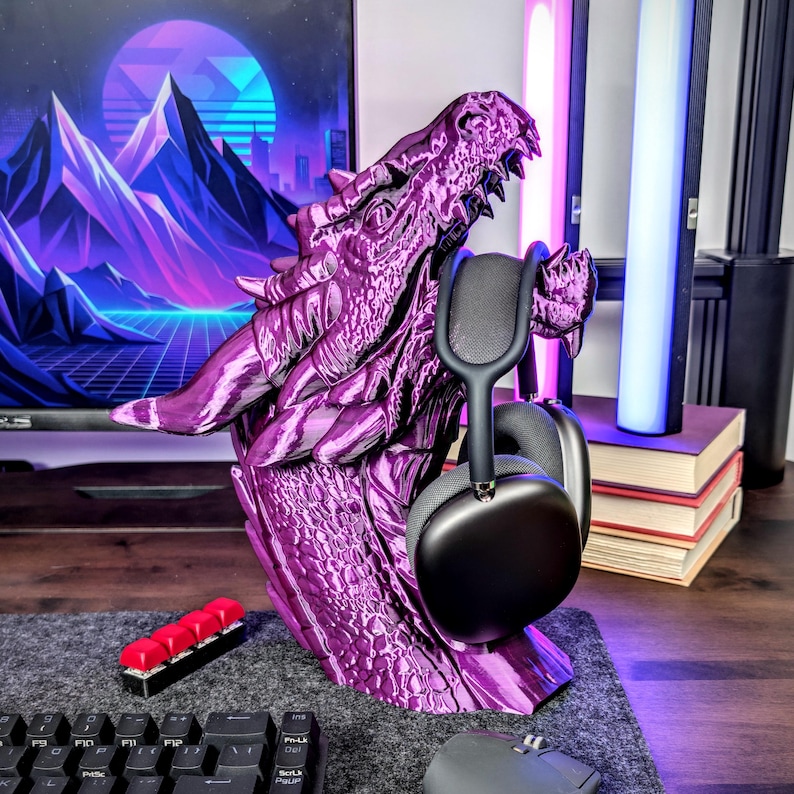 May include: A purple dragon head-shaped headphone stand. The stand is 3D-printed and holds a pair of black headphones. The dragon has detailed scales and a menacing expression. The stand sits on a gray surface.