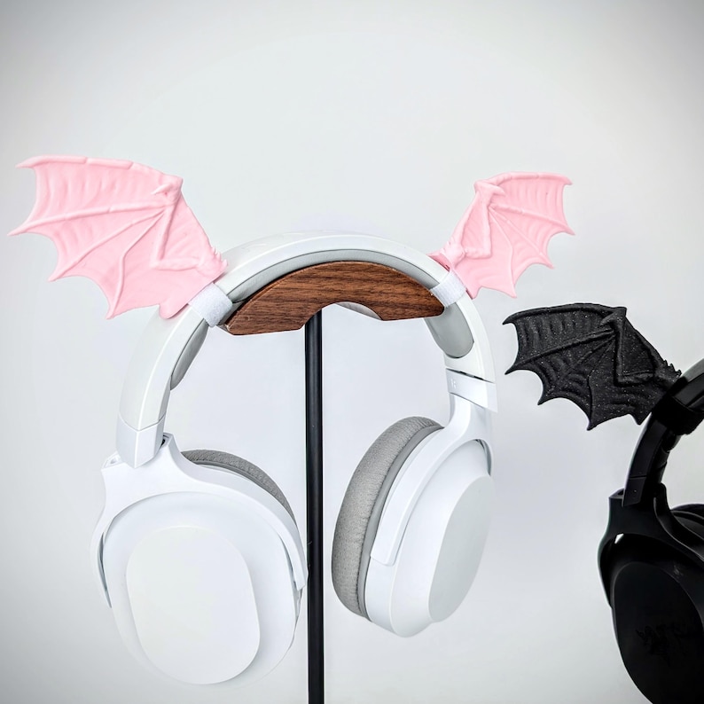 May include: White headphones with gray earcups and pink bat wing attachments are displayed on a black stand. A second set of black headphones with black bat wings is also visible. The headphones have a wooden support.