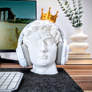 May include: A white plaster bust of a man wearing a gold crown and white headphones. The bust is sitting on a black surface.