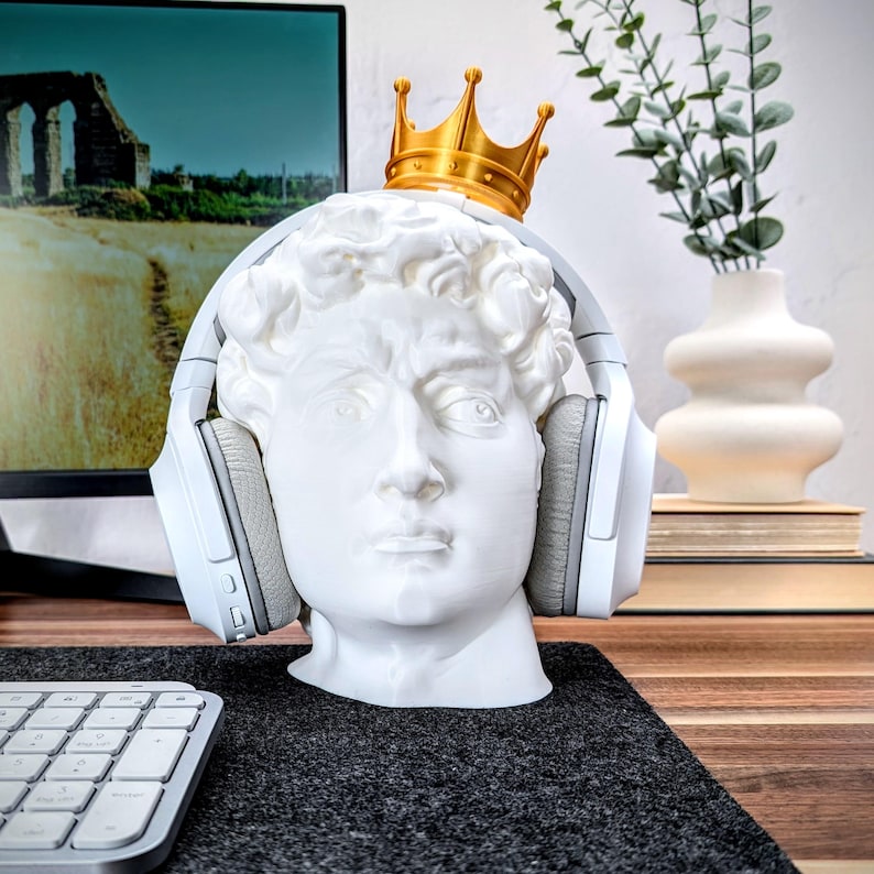 May include: A white plaster bust of a man wearing a gold crown and white headphones. The bust is sitting on a black surface.