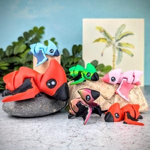 May include: A collection of six small, colorful, hand-painted dinosaur figurines. The dinosaurs are made of polymer clay and have a variety of poses and expressions. The dinosaurs are arranged on a rock surface.