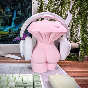 May include: A pink mushroom-shaped headphone stand with white headphones resting on the sides. The stand is on a wooden desk with a keyboard and mouse.