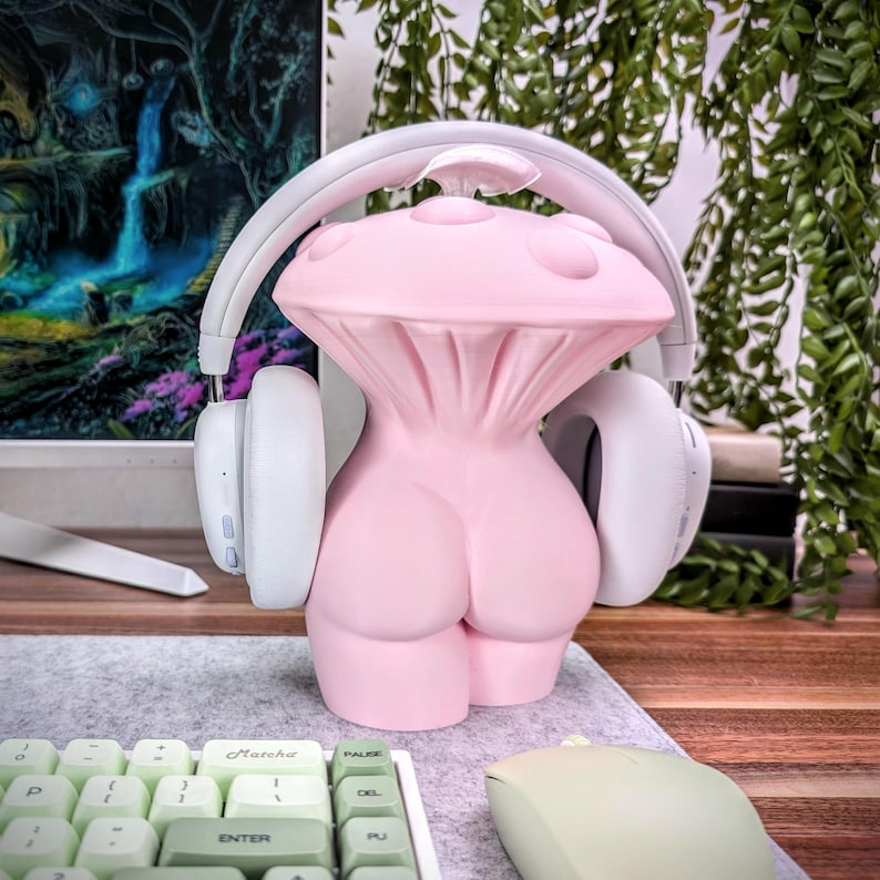 May include: A pink mushroom-shaped headphone stand with white headphones resting on the sides. The stand is on a wooden desk with a keyboard and mouse.