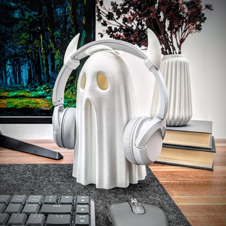 May include: A white ghost figurine with devil horns wearing white headphones. The ghost is sitting on a desk with a keyboard and mouse.