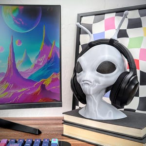 May include: A gray alien bust with large, black headphones resting on its head. The bust is sitting on a stack of books with a black and white checkered background.