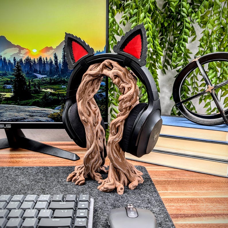 May include: A brown, tree-shaped headphone stand with cat ears. The stand is made of 3D printed plastic and has a natural wood finish. The stand is holding a pair of black headphones.