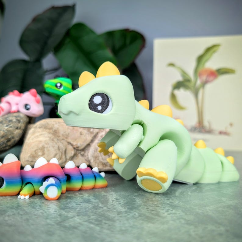 May include: A collection of colorful, 3D-printed dinosaur figurines. The main figurine is light green with yellow accents. Other dinosaurs are pink, green, and rainbow-colored. The figurines are on a gray surface with a blurred background.
