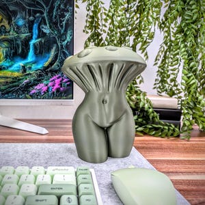 May include: A 3D printed sculpture of a human torso with a mushroom-shaped top. The sculpture is a muted green color and is sitting on a desk.