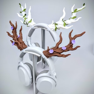 May include: White headphones with decorative antler-shaped attachments. One set of antlers is brown with purple flowers, the other is white with green leaves. The headphones are displayed on a white stand, creating a unique and whimsical aesthetic.