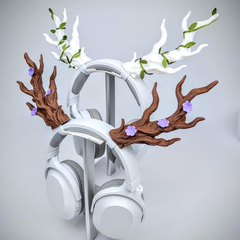 May include: White headphones with decorative antler-shaped attachments. One set of antlers is brown with purple flowers, the other is white with green leaves. The headphones are displayed on a white stand, creating a unique and whimsical aesthetic.