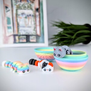 May include: A collection of colorful cat-shaped toys. One is a segmented caterpillar design, another is a calico cat, and a third is a gray cat resting in a rainbow-colored bowl. The toys are on a white surface.