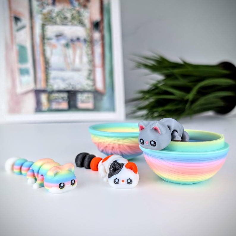 May include: A collection of colorful cat-shaped toys. One is a segmented caterpillar design, another is a calico cat, and a third is a gray cat resting in a rainbow-colored bowl. The toys are on a white surface.