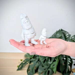 May include: Two white stone statues of a person with a large head and small body. The statues are standing on a person's hand. The larger statue is about twice the size of the smaller statue.