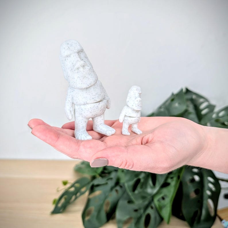 May include: Two white stone statues of a person with a large head and small body. The statues are standing on a person's hand. The larger statue is about twice the size of the smaller statue.