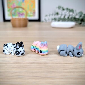 May include: Three small animal figurines: a Dalmatian-style dog, a rainbow-coloured bunny, and a grey bunny. The dog is white with black spots. The rainbow bunny has pastel colours. The grey bunny has a white tail and pink ears.