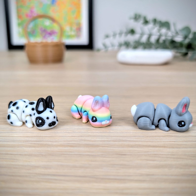 May include: Three small animal figurines: a Dalmatian-style dog, a rainbow-coloured bunny, and a grey bunny. The dog is white with black spots. The rainbow bunny has pastel colours. The grey bunny has a white tail and pink ears.