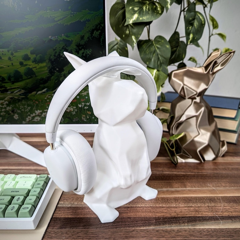 May include: White rabbit-shaped headphone stand with a pair of white over-ear headphones. The stand is on a wooden surface next to a gold origami rabbit.