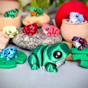 May include: A collection of colorful, 3D-printed frog figurines. The frogs are in various colors, including green, blue, red, pink, and purple. Some frogs are on lily pads or rocks. The frogs have large, expressive eyes and are detailed.