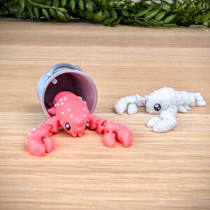 May include: Two small lobster figurines, one pink with white spots and one white with black spots. The pink lobster is in a small grey bucket.