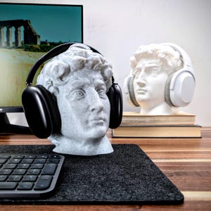 May include: A white marble bust of a man wearing black over-ear headphones. The bust is sitting on a black desk mat with a keyboard in the foreground.