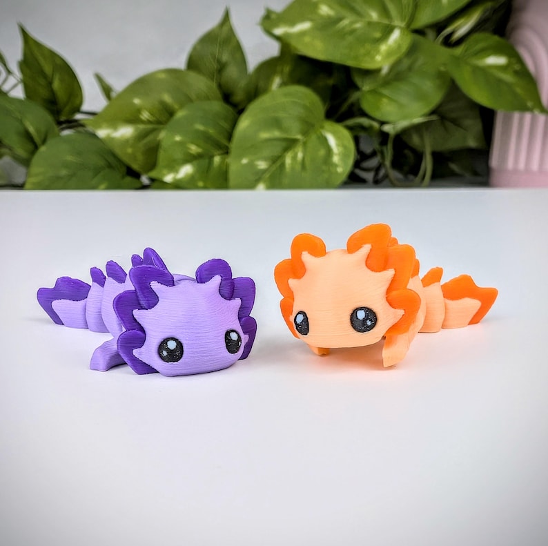 May include: Two small, 3D printed axolotl figurines. One is purple with a textured finish and the other is orange with a smooth finish.