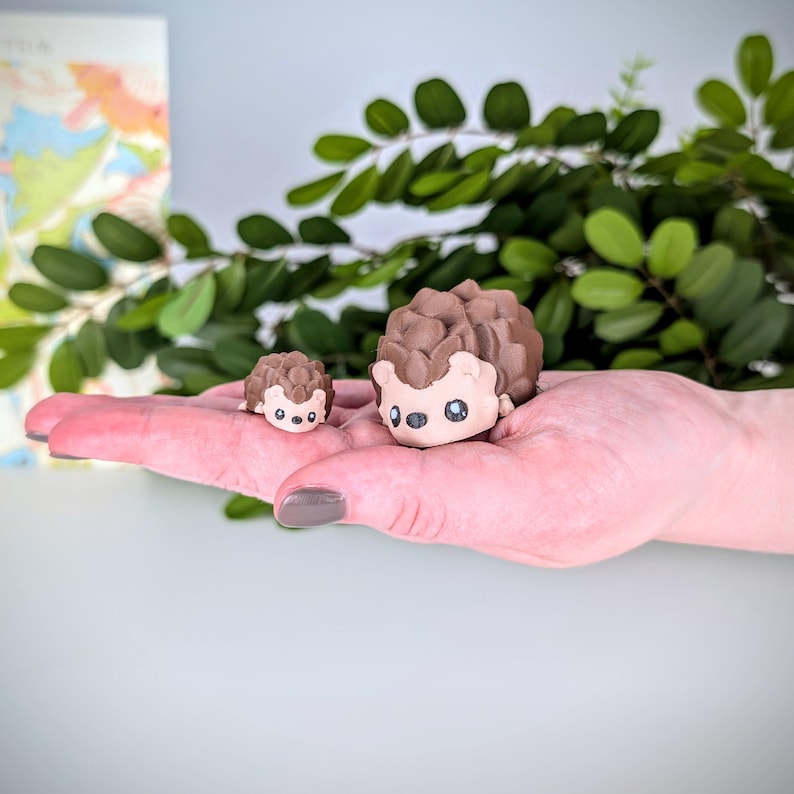 May include: Two brown and black ceramic hedgehogs, one larger than the other, are sitting on a hand. The hedgehogs have small black eyes and white noses.