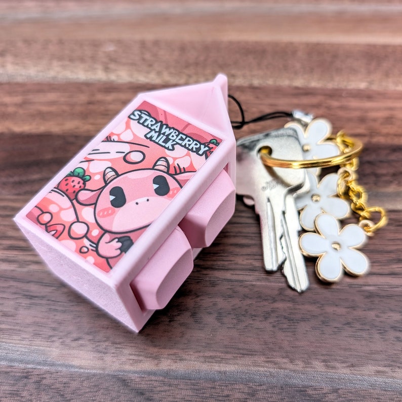 May include: A pink plastic milk carton keychain with a cartoon cow and strawberry design. The keychain says "Strawberry Milk" and has a gold key ring with white flower charms.